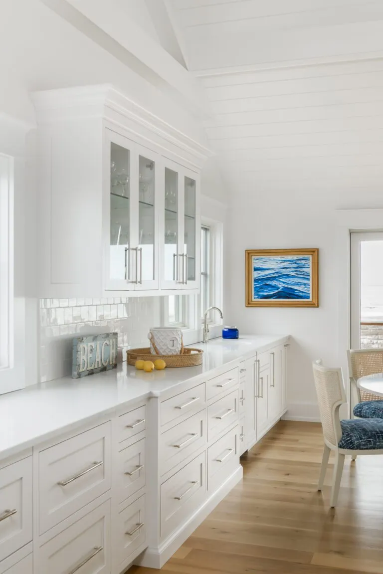 Spacious coastal kitchen featuring white cabinets, glass-front upper cabinets, a long countertop, and ocean-themed decor, including a "BEACH" sign and a seascape painting.