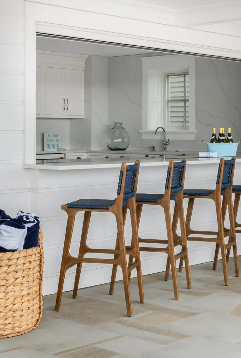 Outdoor kitchen bar with white countertops, wooden bar stools with blue seats, a wicker basket with rolled towels, and a view into a modern indoor kitchen.