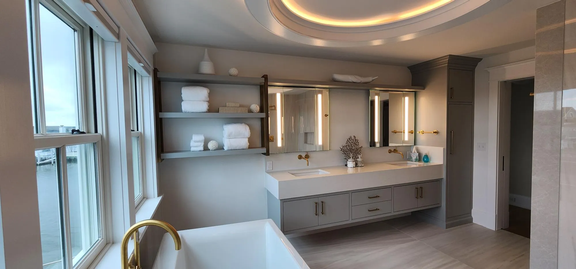 Modern bathroom featuring a large bathtub, double vanity with gold fixtures, and shelves stocked with towels, overlooking an ocean view.