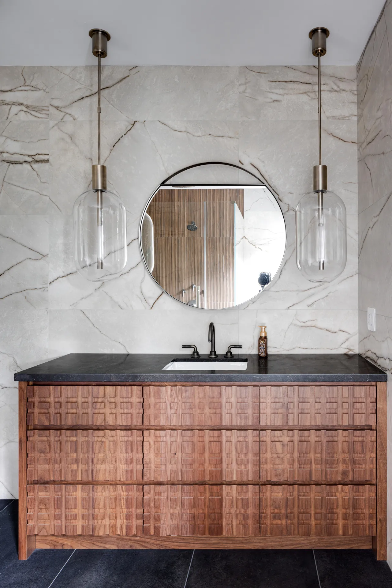 Bathroom featuring a rustic textured wood vanity, black countertop, round mirror, and pendant lighting.