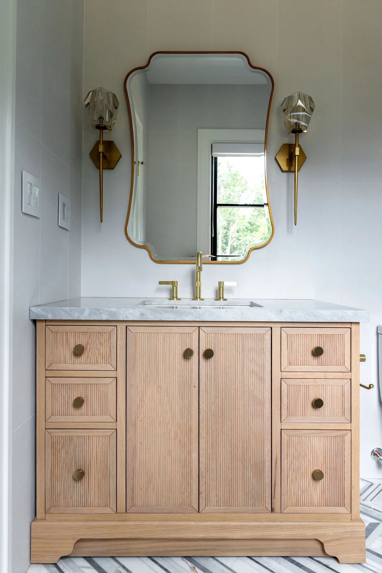 Stylish bathroom featuring a light wood vanity, marble countertop, brass fixtures, and a decorative mirror.