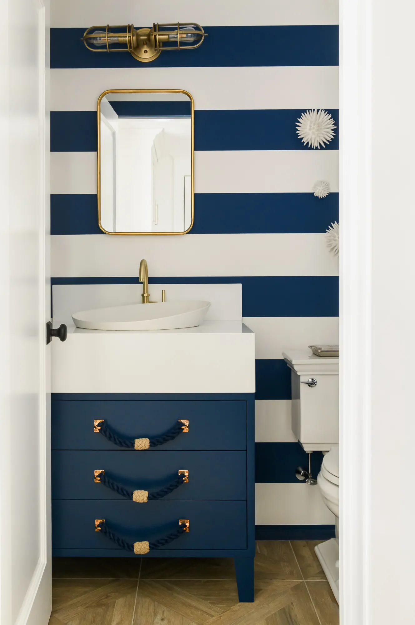 Nautical-themed bathroom featuring blue and white striped walls, blue vanity with gold fixtures, and a unique light fixture.