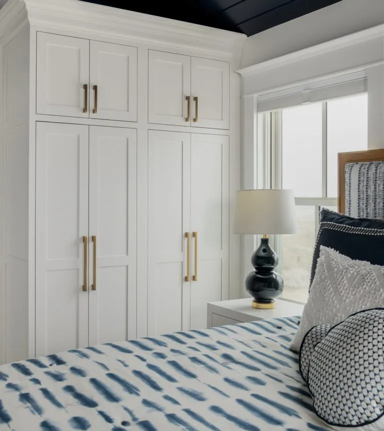 Stylish nautical-inspired bedroom featuring navy blue wallpaper with white rope patterns, a large white wardrobe with gold handles, and a view into a modern bathroom.