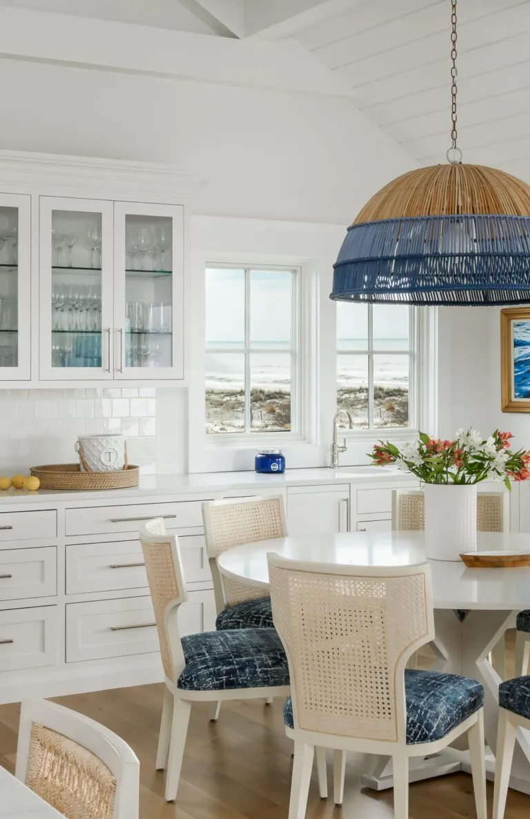 Bright coastal dining room featuring a round white table, wicker chairs with blue cushions, a blue and natural fiber pendant light, and a stunning ocean view.