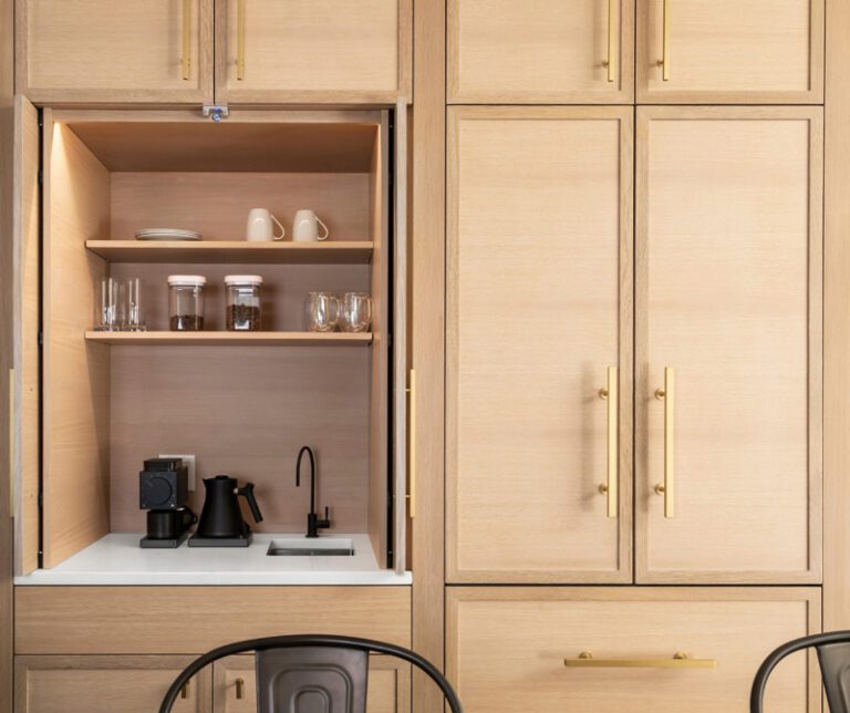 A built-in coffee bar hidden within white oak cabinetry, revealed by open retractable doors, showing shelves, a small sink, faucet, and coffee maker, surrounded by more white oak cabinets with brass hardware.