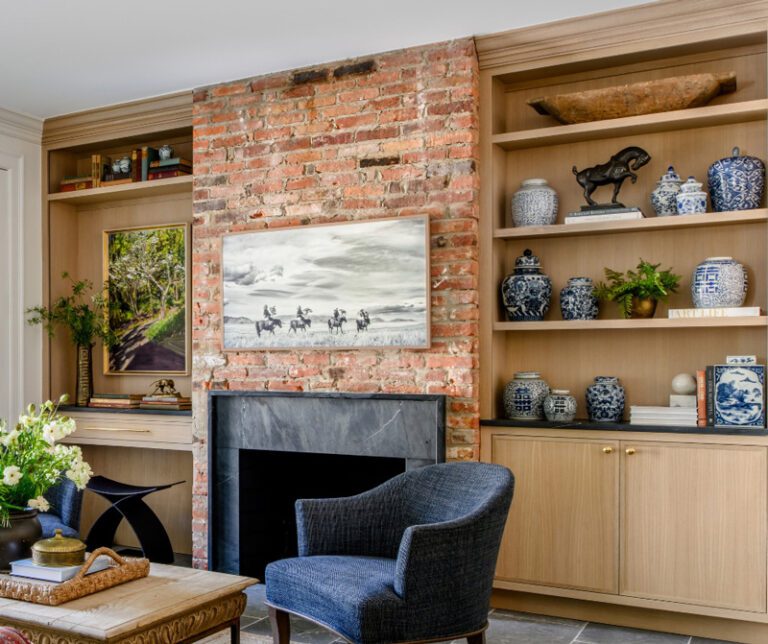 Built-in white oak cabinetry and shelving flanking an exposed brick fireplace with a dark stone surround in a living room, styled with books, pottery, and art.