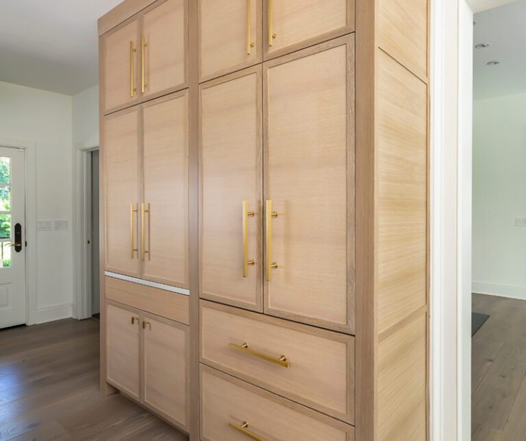 A tall bank of floor-to-ceiling white oak cabinetry with brass hardware, featuring various door and drawer sizes, likely functioning as a pantry or integrated storage unit.