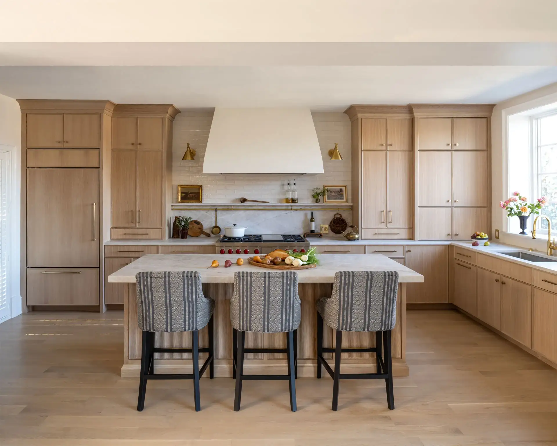 A spacious luxury kitchen featuring custom white oak cabinetry throughout, integrated refrigerator towers, a large island, a professional range, subtle tile backsplash, and brass hardware.