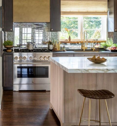 A luxury kitchen island featuring reeded white oak cabinetry, a marble countertop, and a leather bar stool with brass legs, with dark cabinets and a range in the background.