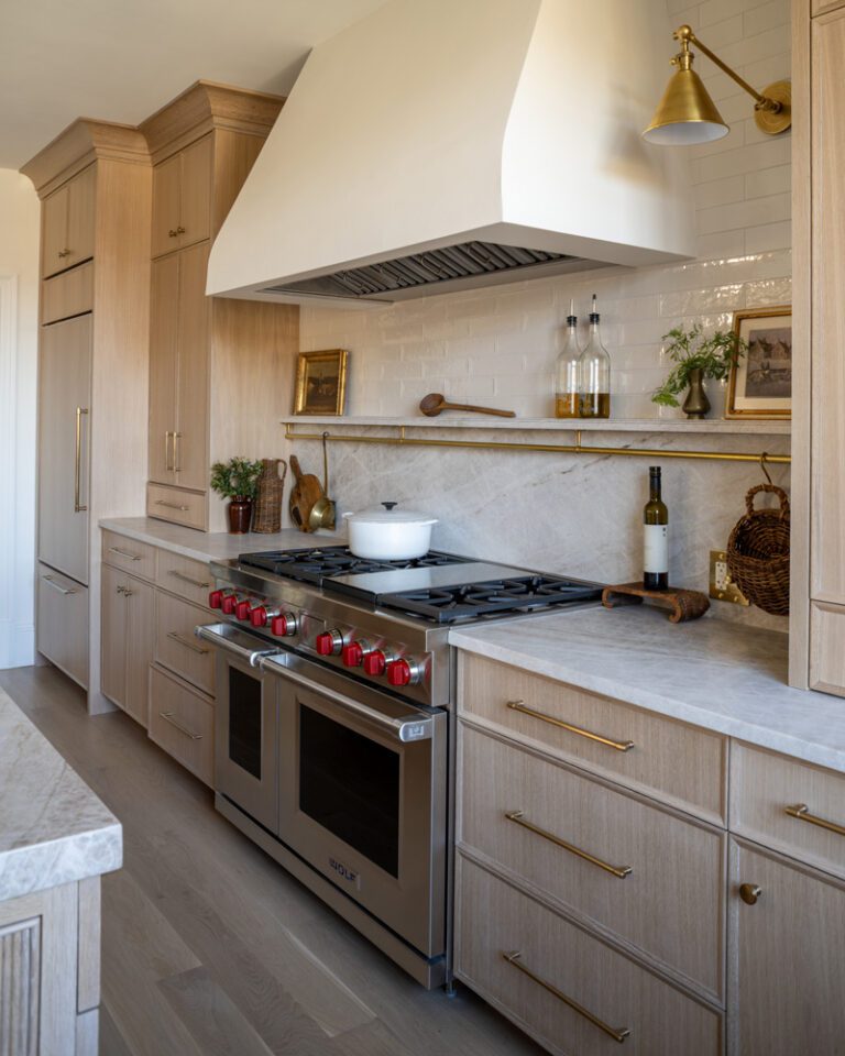 A luxury kitchen featuring timeless white oak cabinetry, a professional stainless steel range, marble backsplash and countertop, and brass hardware and wall sconce.