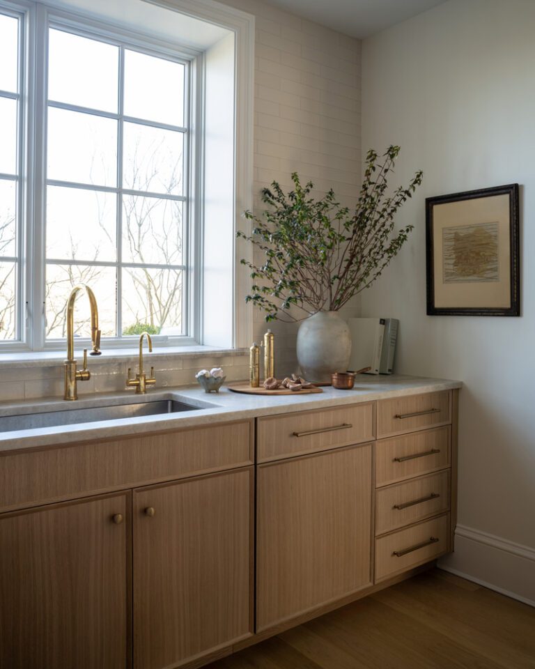 A kitchen or service area sink with light-finished white oak cabinetry below, light stone countertop, brass faucet and hardware, and a large window.