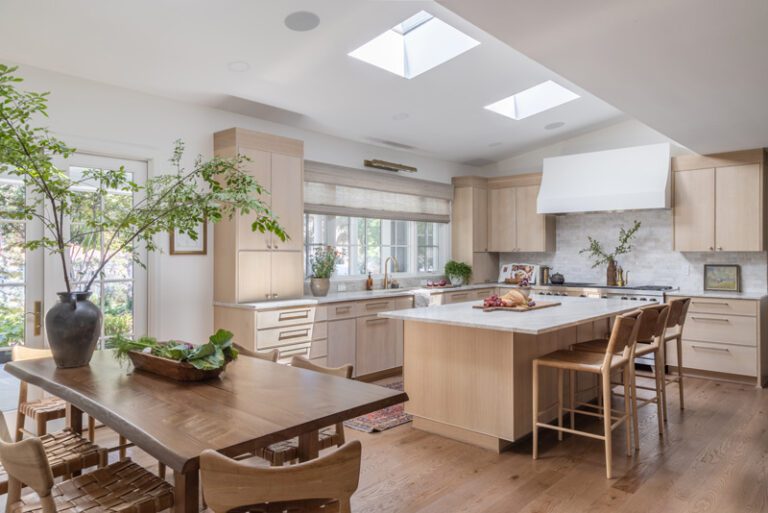 A bright, spacious kitchen and dining area featuring light-finished white oak cabinetry, a large island, a wooden dining table and chairs, skylights, and large glass doors.
