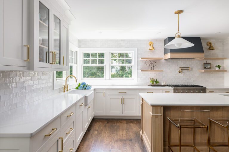 A transitional kitchen featuring a white oak island, white perimeter cabinets, white subway tile backsplash, a black and wood range hood, floating white oak shelves, brass hardware, and clear bar stools.
