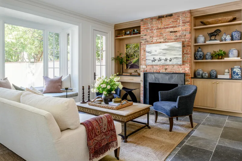 A quiet luxury living room features a white sofa, a bay window seat, an exposed brick fireplace with a television, built-in light wood shelving, and grey floor tiles.