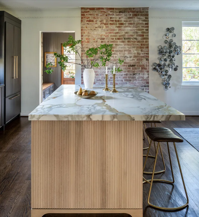 A quiet luxury kitchen features a grand island with a veined marble countertop, fluted wood paneling, dark stools, and an exposed brick accent wall.