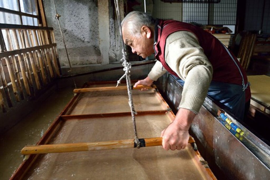 An elderly Japanese craftsman carefully making traditional washi paper, holding a large wooden screen frame over a vat of water and pulp in his workshop.