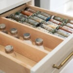 A well-organized kitchen drawer with custom light wood inserts neatly holding rows of spice jars. The off-white cabinet has a sleek brass pull handle, showcasing Japandi-style organization.