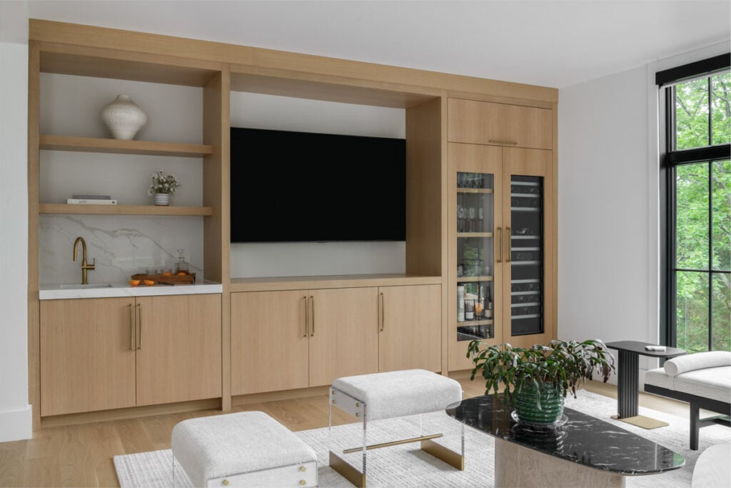 A custom light oak entertainment center and wet bar in a Japandi-style living room, featuring a built-in TV, wine fridge, and marble backsplash. Minimalist furniture sits in the foreground.