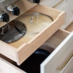 A custom kitchen cabinet drawer solution under a Wolf range for storing pots and pans. A shallow sliding wooden tray holds lids above a deeper compartment for cookware.