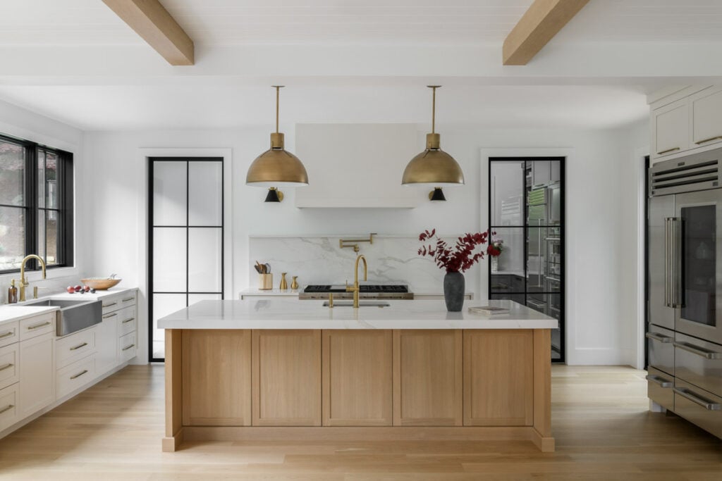 A bright and airy Japandi-style luxury kitchen with a large light oak island, white perimeter cabinets, marble-look quartz countertops, and oversized brass dome pendant lights.
