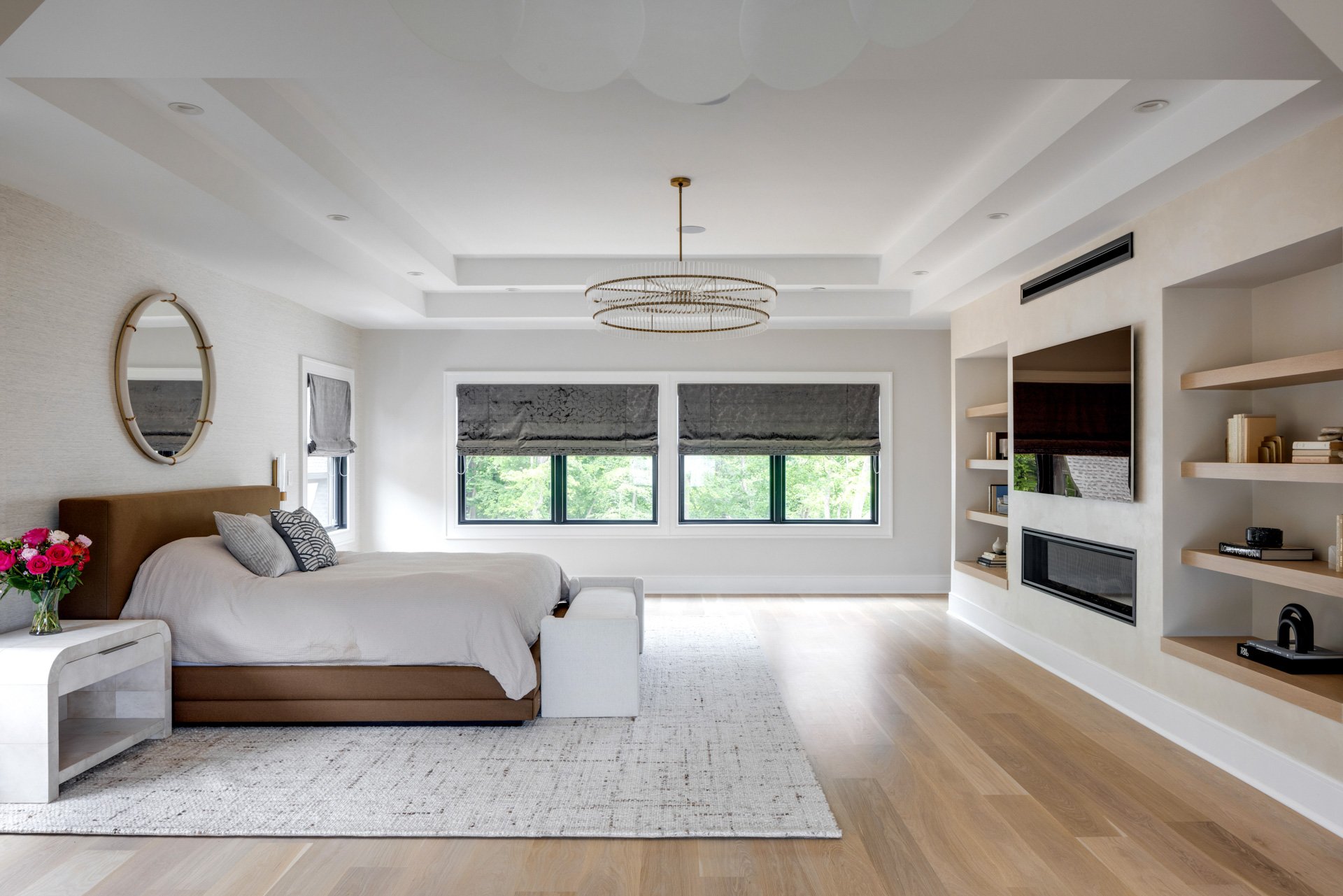 A wide and a detail shot of a spacious Japandi primary bedroom with light oak floors, a tray ceiling, and a custom media wall featuring a linear fireplace and curated floating oak shelves.