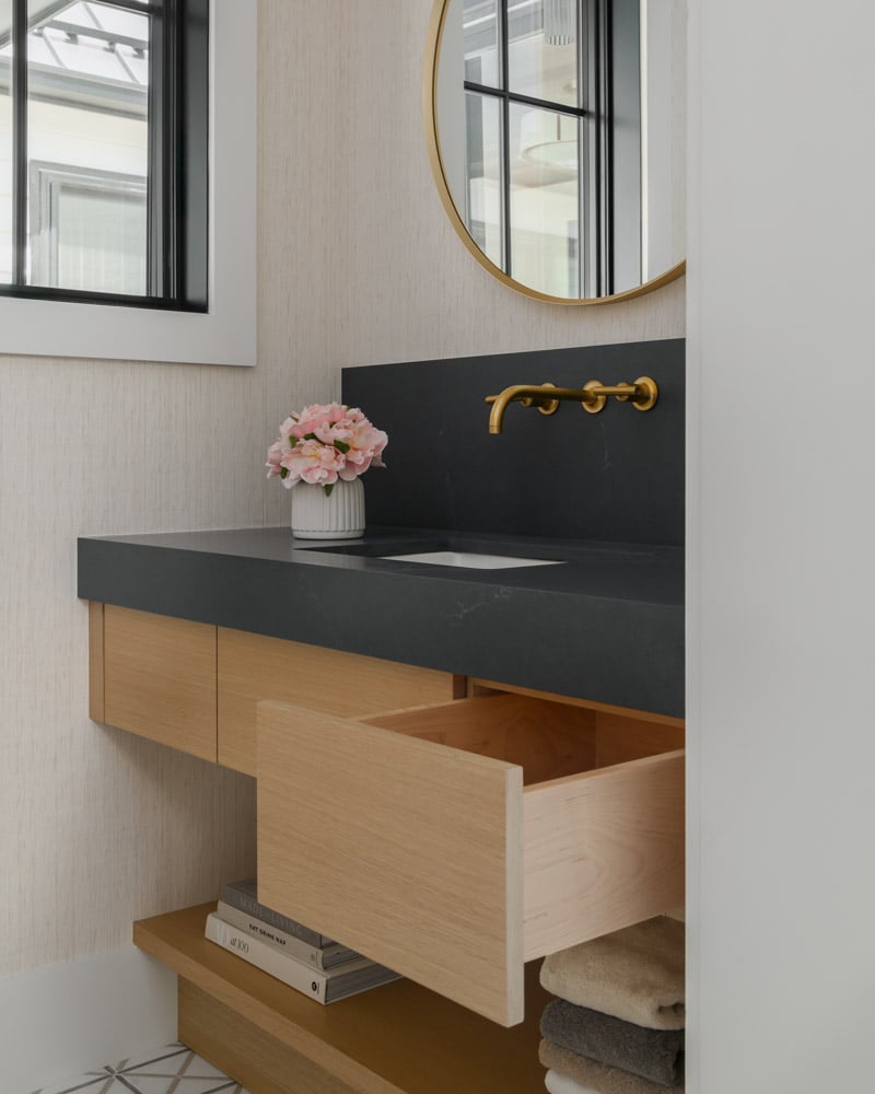 A Japandi-style bathroom vanity with a light oak floating cabinet, a dark honed stone countertop with an integrated sink, and a wall-mounted brushed brass faucet. A round gold-framed mirror hangs above.