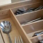A custom light wood cutlery organizer inside a kitchen drawer. The silver forks and spoons are neatly arranged in compartments. The Master Design Cabinetry logo is visible inside the drawer.