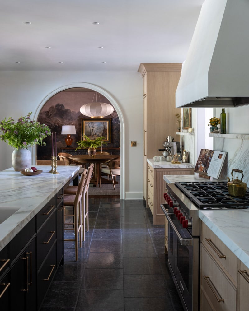 A luxury kitchen with dark stone floors and a dark island with a marble top, leading through a large archway to a moody dining room with a landscape mural.