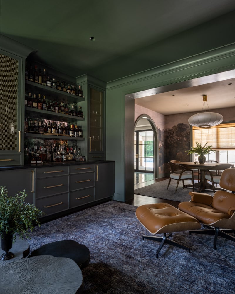 A view from a luxurious lounge area, showing a tan leather Eames chair in the foreground and a sophisticated, custom-built home bar in deep green with brass mesh cabinet doors and a view into the adjacent dining room.