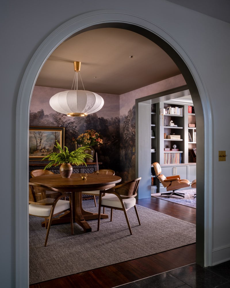 View through an elegant archway into a dining room with a dramatic, moody landscape mural. A modern wood dining table is surrounded by caned chairs, and a large, white paper lantern pendant hangs above, exemplifying quiet luxury.