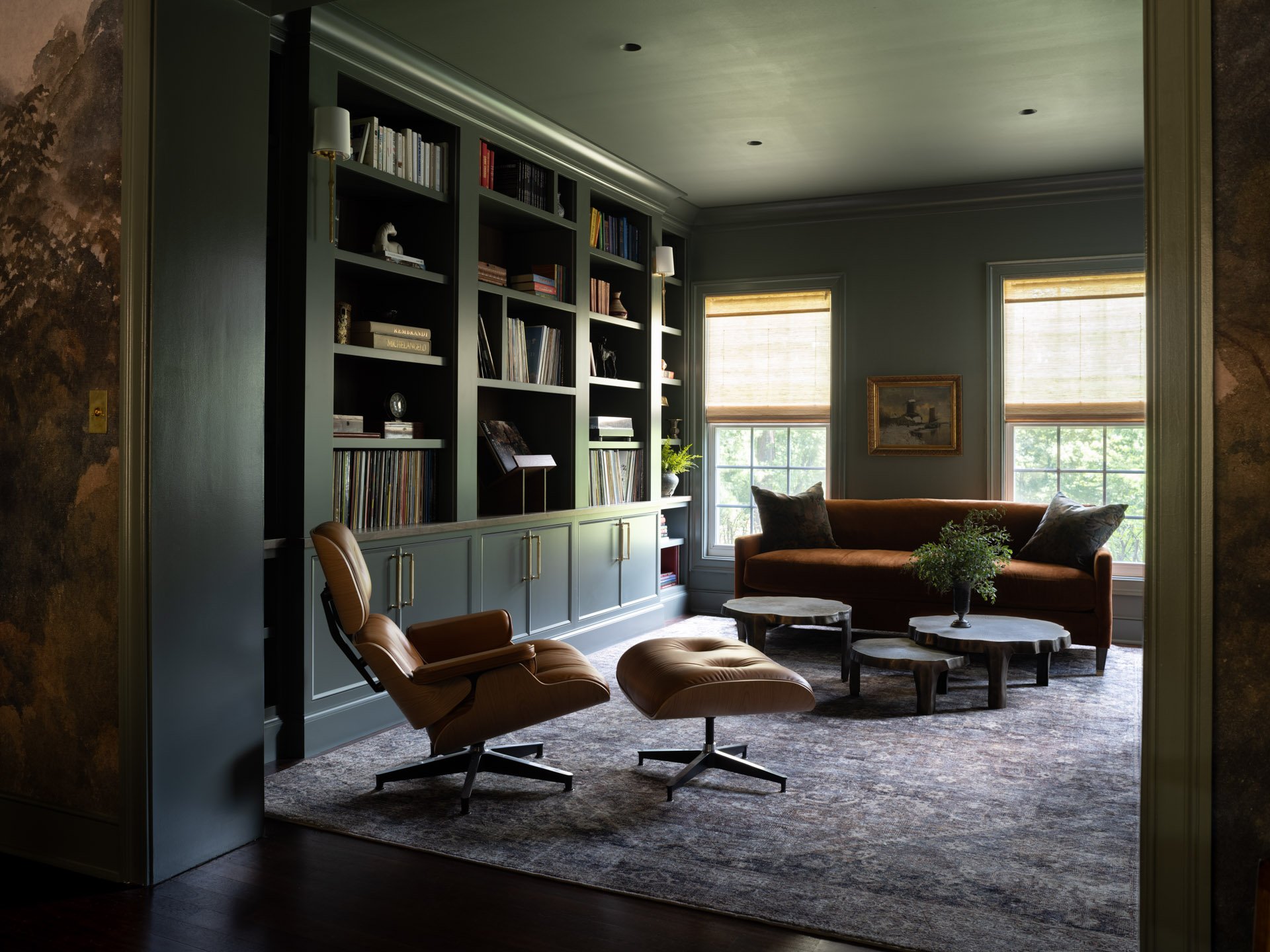 A wider view of a sophisticated music and reading room with deep green walls and built-in bookshelves. A classic tan leather Eames lounge chair and ottoman sit in the foreground, facing an orange sofa and windows with natural light.