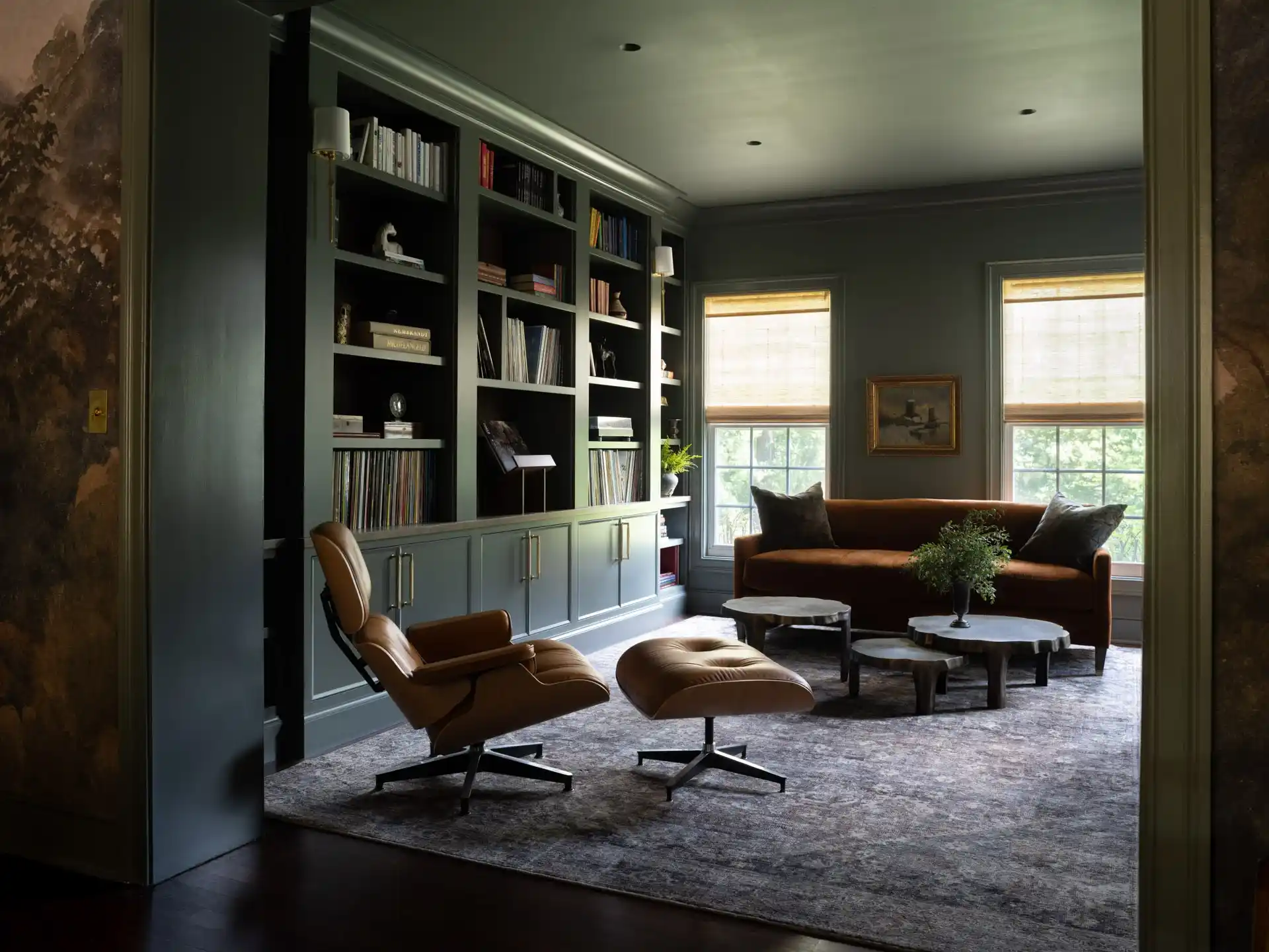 A wider view of a sophisticated music and reading room with deep green walls and built-in bookshelves. A classic tan leather Eames lounge chair and ottoman sit in the foreground, facing an orange sofa and windows with natural light.