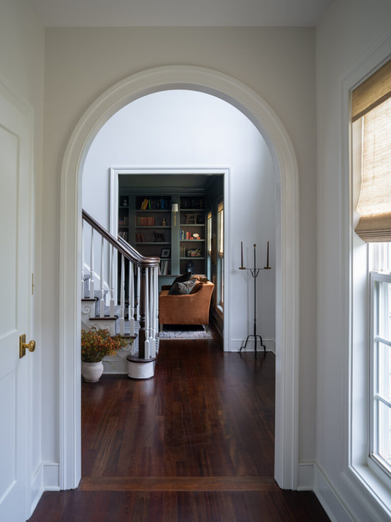 A view down a bright hallway with dark wood floors, looking through an elegant white archway into a moody, dark green library. A classic staircase is visible on the left.