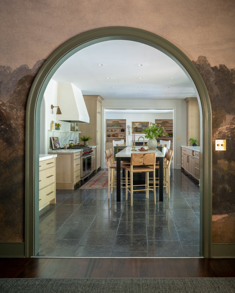 A stunning view through a green-trimmed archway into a luxury kitchen with light oak cabinets, a marble island, and dark slate floors. The foreground wall features a rich, textured mural.