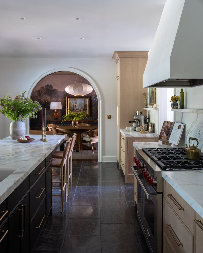 A close-up view of a high-end kitchen featuring a dark island with a thick marble countertop, a Wolf gas range, and light wood cabinetry. An archway in the background reveals the adjoining dining room.
