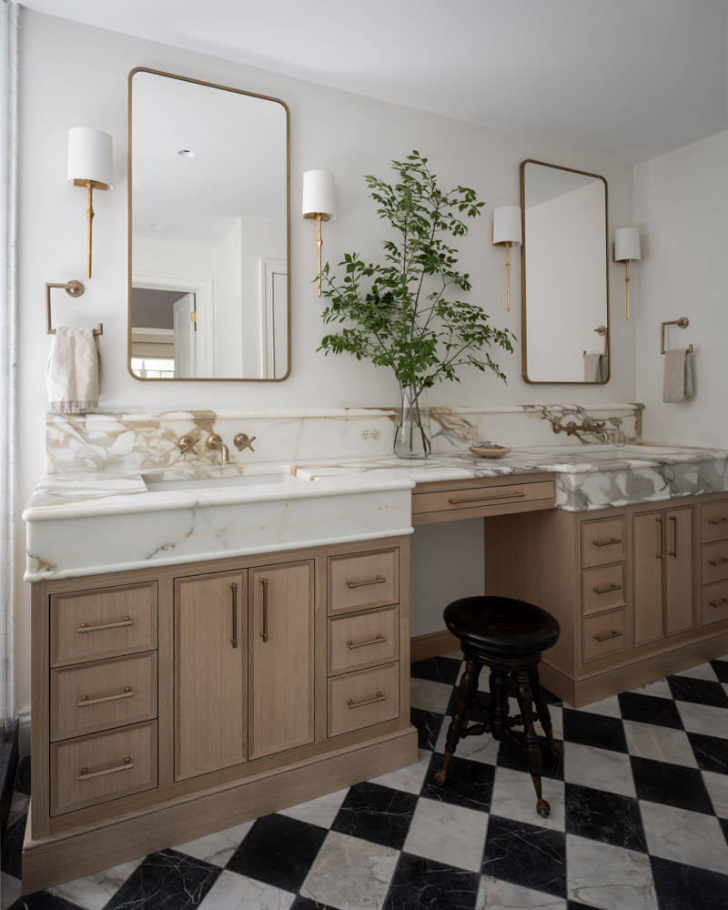 A luxurious primary bathroom with a black and white checkered marble floor, dual light oak vanities with thick Calacatta Gold marble countertops, and elegant brass mirrors and sconces.