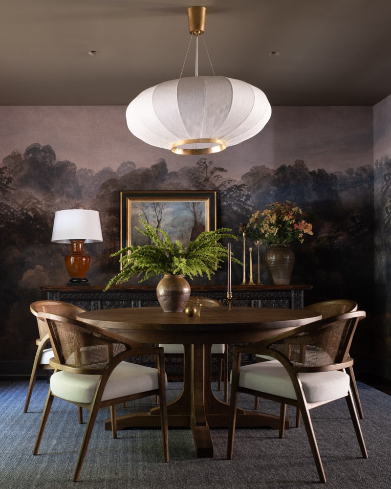 A moody and atmospheric Japandi-influenced dining room featuring a dark landscape wall mural, a large sculptural paper lantern, a round wood table, and caned chairs.