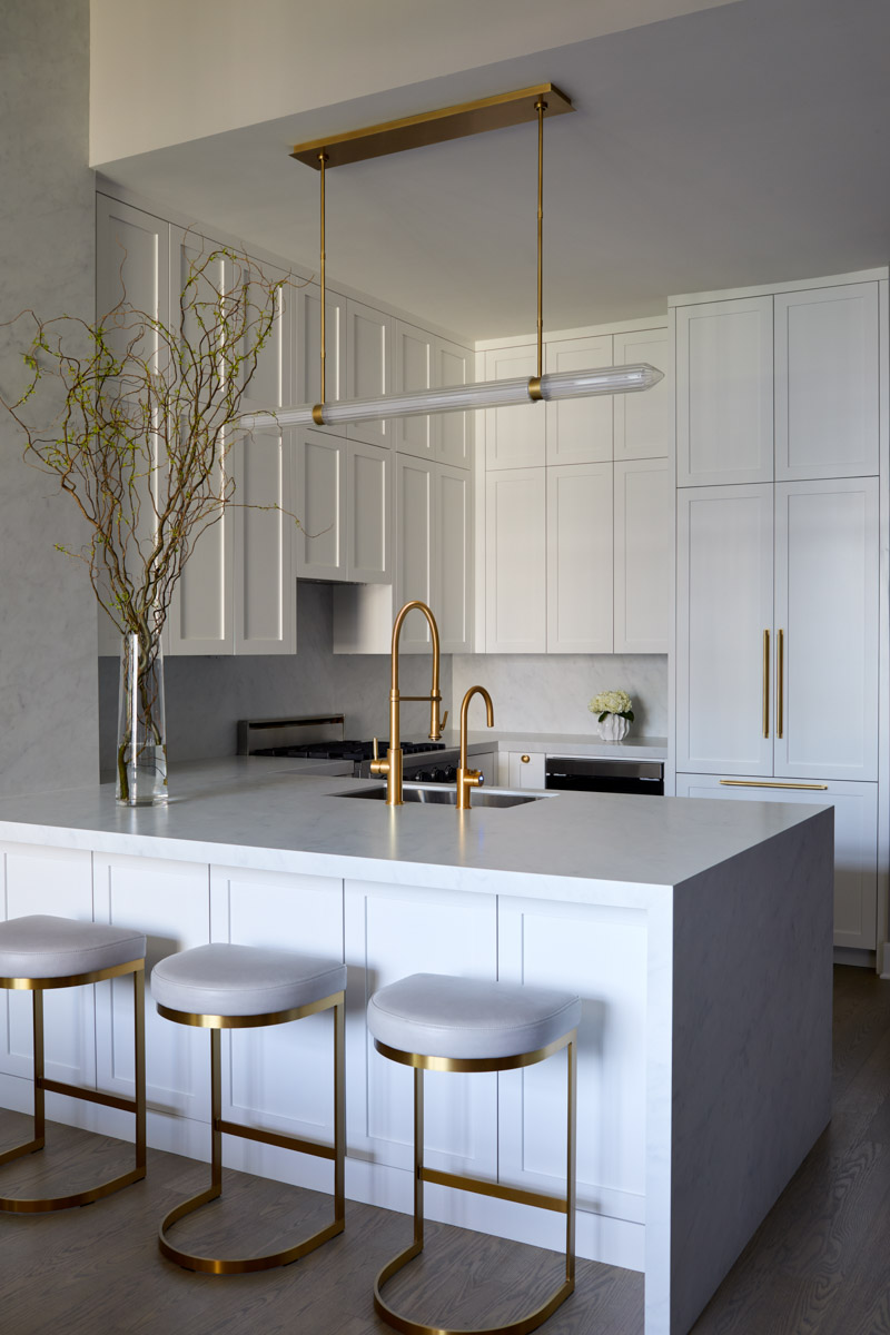 A sleek and modern all-white minimalist kitchen featuring handleless cabinetry, marble-look countertops and backsplash, a statement linear brass pendant light, and a tall brass faucet.